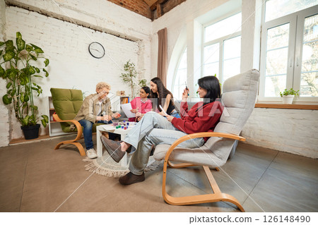 Woman relaxes in armchair while friends paint at table in bright room, observing activity. Concept of leisure, relaxation, friends, observation, indoor life. 126148490