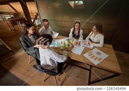 Business professionals review charts and documents, shares ideas, sits at large table. Concept of meeting, analysis, strategy, teamwork, collaboration. Business professionals review charts and documents, shares ideas, sits at large table. Concept of meeting, analysis, strategy, teamwork, collaboration. 126148494