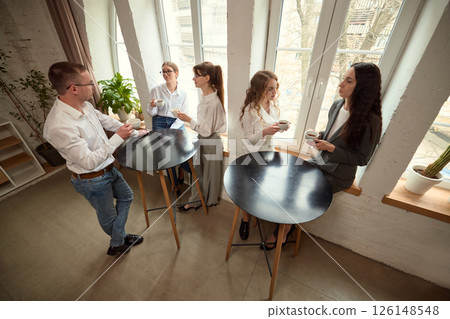 Group of colleagues stand around high tables in modern office, talk and drink coffee during break. 126148548