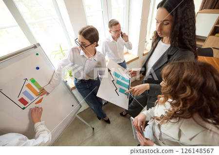 Business team discusses charts and data near whiteboard in modern office, pointing at visual aids. 126148567