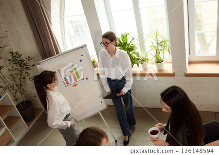 Colleague points to chart on whiteboard, presents to team members in bright office, explaining data visuals. Colleague points to chart on whiteboard, presents to team members in bright office, explaining data visuals. 126148594