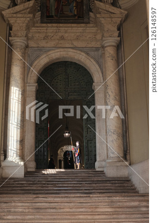 Swiss Guard in traditional Renaissance uniform holding ceremonial halberd under arched marble doorway inside Vatican City with dramatic light and historic architecture guarding entrance to sacred area Swiss Guard in traditional Renaissance uniform holding ceremonial halberd under arched marble doorway inside Vatican City with dramatic light and historic architecture guarding entrance to sacred area 126148597