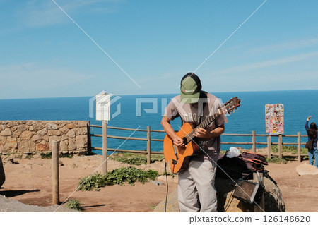 Street musician playing acoustic guitar at scenic coastal viewpoint in Cabo da Roca Sintra Portugal with ocean and stone wall background on sunny day. High quality photo 126148620
