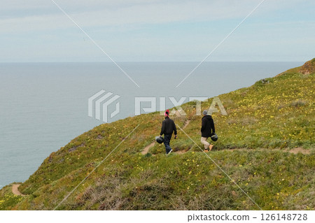 Hikers walking along scenic coastal trail at Cabo da Roca Sintra Portugal with grassy cliffs overlooking Atlantic Ocean under cloudy sky westernmost mainland Europe outdoor travel landscape. High 126148728