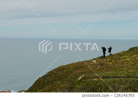 Hikers walking along scenic coastal trail at Cabo da Roca Sintra Portugal with grassy cliffs overlooking Atlantic Ocean under cloudy sky westernmost mainland Europe outdoor travel landscape. High 126148765