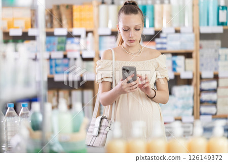 Young pregnant woman scrolling phone while looking for something in drugstore Young pregnant woman scrolling phone while looking for something in drugstore 126149372