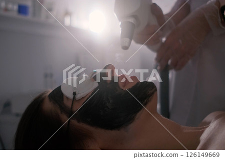 A woman is undergoing a carbon laser facial treatment. Her face is covered with a black carbon mask, protective goggles shield her eyes. 126149669