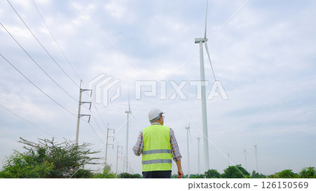 Engineer walking through wind turbine field inspecting renewable energy infrastructure under cloudy sky 126150569