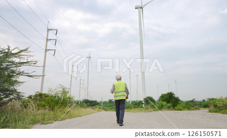 Engineer walking through wind turbine field inspecting renewable energy infrastructure under cloudy sky 126150571