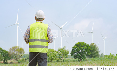 Engineer holding blueprint while surveying wind turbines at renewable energy site under bright sky conditions. 126150577
