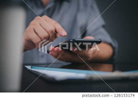 Hand operating cellphone at workplace desk, business lifestyle. Modern office worker using mobile technology, professional environment capture with documents." 126153174