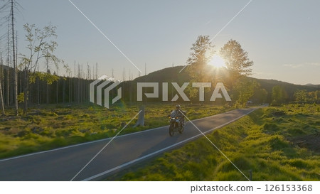 Driver riding motorcycle on empty asphalt road, spring mountains during sunset 126153368