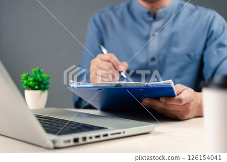 A professional in a blue shirt holding a clipboard and writing, with a laptop and small plant in the background, symbolizing office work, documentation, task management and productivity. 126154041