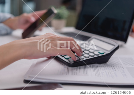 Office worker performing calculations with a calculator and smartphone on the table, analyzing business data or financial reports. Office worker performing calculations with a calculator and smartphone on the table, analyzing business data or financial reports. 126154606