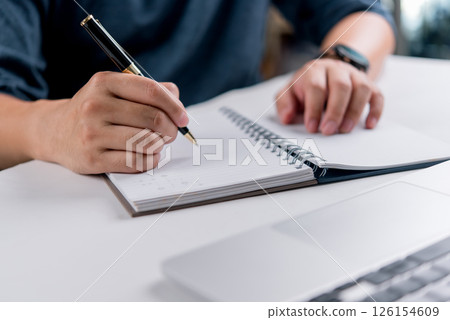 Hand writing on spiral notebook with pen and laptop, concept of education, online learning, planning, productivity, and personal development. Hand writing on spiral notebook with pen and laptop, concept of education, online learning, planning, productivity, and personal development. 126154609