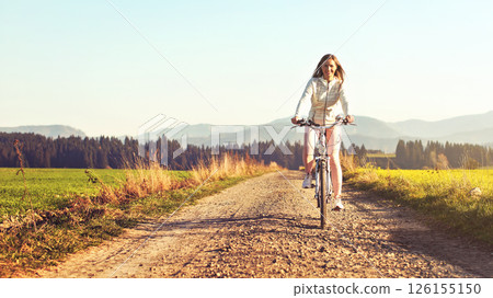 Young woman rides bicycle on dusty country road, afternoon sun shines at her, view from front 126155150