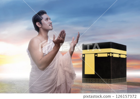 Indonesian southeast asian muslim man wearing white traditional clothes or ihram raises his hand and prays in front of the Kaaba. Concept of hajj or umrah Indonesian southeast asian muslim man wearing white traditional clothes or ihram raises his hand and prays in front of the Kaaba. Concept of hajj or umrah 126157372