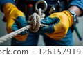 Hands clad in gloves expertly secure a dynamic rope knot while wearing a well-used climbing harness, showcasing the skill and focus required for industrial climbing. 126157919