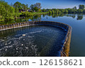 Circular weir overflowing into river on sunny day 126158621