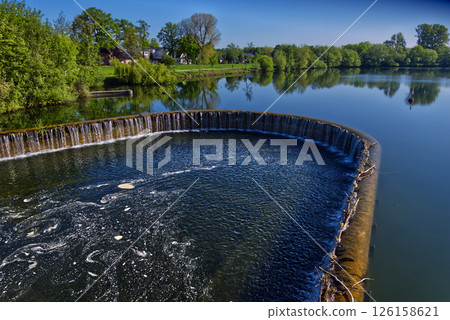 Circular weir overflowing into river on sunny day 126158621