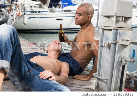 Romantic couple of young men sitting and relaxing together on the pavement in the port Romantic couple of young men sitting and relaxing together on the pavement in the port 126159352