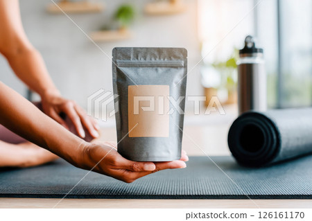 Hand holding a gray packaging bag on a yoga mat with water bottle and fitness accessories in background 126161170