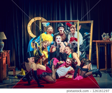 Ensemble of cabaret performers in colorful vintage costumes and dramatic makeup pose theatrically around large gold frame on red carpet. Ensemble of cabaret performers in colorful vintage costumes and dramatic makeup pose theatrically around large gold frame on red carpet. 126161297
