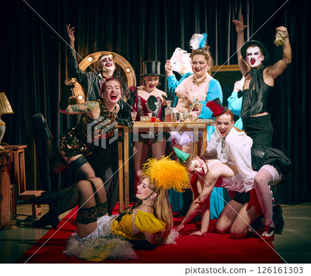 Laughing cabaret troupe gathers around makeup table with drinks, wearing vintage clothes, posing with expressive faces. Laughing cabaret troupe gathers around makeup table with drinks, wearing vintage clothes, posing with expressive faces. 126161303
