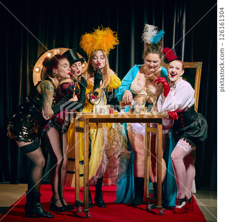 A group of female cabaret performers in diverse vintage costumes gather around a small table laden with makeup and props on a red carpet. A group of female cabaret performers in diverse vintage costumes gather around a small table laden with makeup and props on a red carpet. 126161304