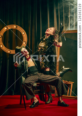 Expressive laughter and dramatic poses of two cabaret performers in vintage costumes on confetti-covered red carpet with smoke, lively moment. Expressive laughter and dramatic poses of two cabaret performers in vintage costumes on confetti-covered red carpet with smoke, lively moment. 126161319