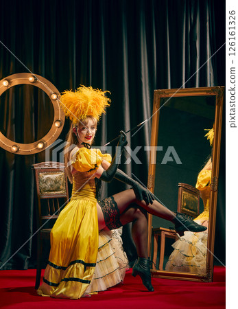 Female elegant performer, dressed in yellow vintage style outfit, seated elegantly on chair on red carpet, posing with long cigarette holder. Female elegant performer, dressed in yellow vintage style outfit, seated elegantly on chair on red carpet, posing with long cigarette holder. 126161326