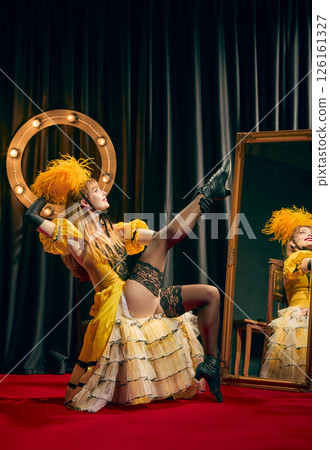 Female cabaret performer in vibrant yellow vintage dress strikes dynamic pose, reflection in large mirror, against backdrop of confetti. Female cabaret performer in vibrant yellow vintage dress strikes dynamic pose, reflection in large mirror, against backdrop of confetti. 126161327