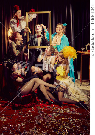 Group of expressive cabaret artists, adorned in vintage clothing, playfully interact with gold frame, posing on confetti-covered red carpet. 126161343