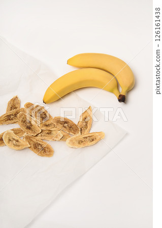 Two fresh bananas, next to which are dried banana slices on parchment paper. White background. 126161438