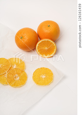 Two fresh oranges, next to which are dried orange slices on parchment paper. White background. 126161439