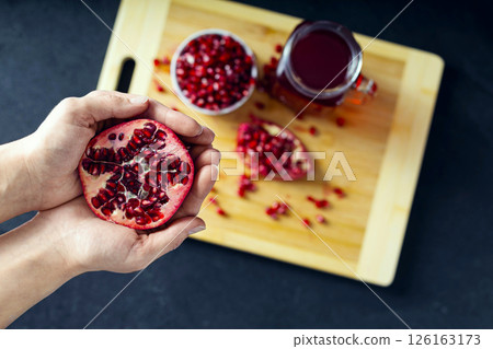 Hands holding cutted pomegranate and a ceramic bowl full of pomegranate seeds, cutted pomegranate and a glass of pomegranate juice on the background lie on the wooden cutting board. View from the top Hands holding cutted pomegranate and a ceramic bowl full of pomegranate seeds, cutted pomegranate and a glass of pomegranate juice on the background lie on the wooden cutting board. View from the top 126163173