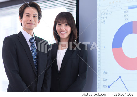 A man and a woman in suits standing in front of a screen in a conference room 126164102