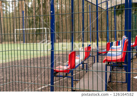 Empty red and white plastic chairs on sidelines of soccer field behind fence Empty red and white plastic chairs on sidelines of soccer field behind fence 126164582