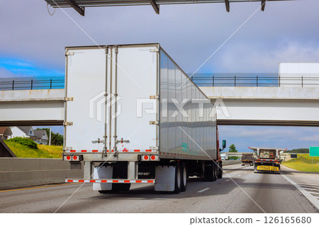 Large trucks travel along busy American highway with an overpass in suburban setting under cloudy sky. 126165680