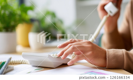 Person communicating on a landline phone at a desk filled with plants and office supplies during daylight hours 126166334