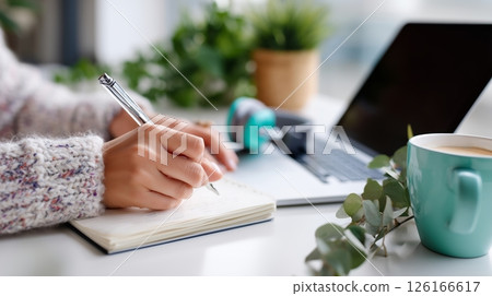 Person taking notes at a home workspace with plants and a laptop during daytime Person taking notes at a home workspace with plants and a laptop during daytime 126166617