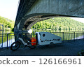 Electric bicycle towing small caravan under bridge near river and green hill 126166961