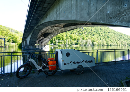 Electric bicycle towing small caravan under bridge near river and green hill 126166961