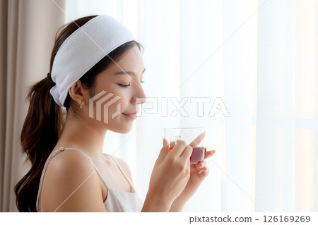 Beautiful young asian woman with headband enjoying tea standing near window with satisfied. 126169269