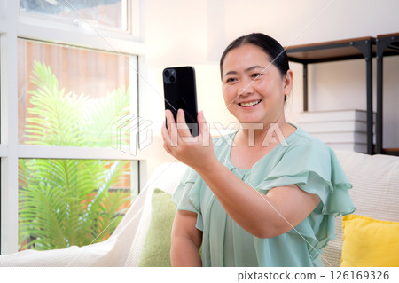 Asian senior woman smiling video call with smartphone in cozy living room at home. 126169326