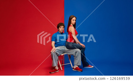 Serious young man and woman, couple sitting together on stool with neutral expressions, against vivid red and blue background. 126169539