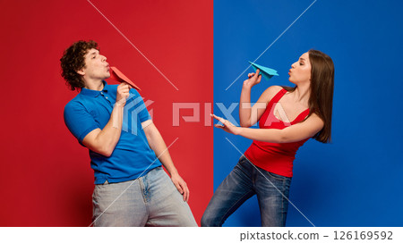 Full-length of young man and woman, couple playfully holding paper airplanes, looking at each other with playful expressions Full-length of young man and woman, couple playfully holding paper airplanes, looking at each other with playful expressions 126169592