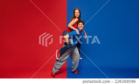 Laughing woman rides on mans back as both pose with energy and joy on red blue split background. Couple in casual clothes. Laughing woman rides on mans back as both pose with energy and joy on red blue split background. Couple in casual clothes. 126169596