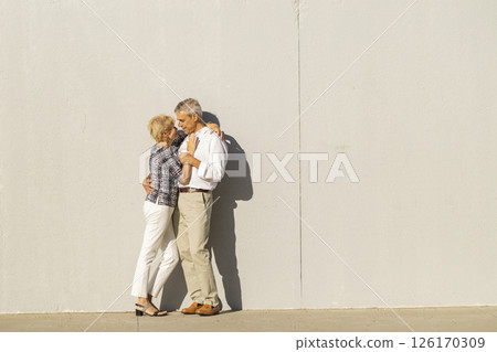 Senior couple embracing tenderly against a wall in sunlight Senior couple embracing tenderly against a wall in sunlight 126170309