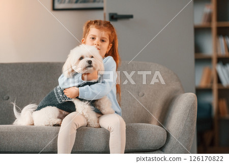 Front view. On the sofa. Little girl is with maltese dog indoors in domestic room 126170822